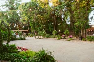 A peaceful garden courtyard with a gravel path, lush greenery, flowering plants, and rustic wooden buildings in the background.
