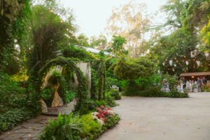 Enchanting garden pathway with floral arches and lush greenery leads to an outdoor courtyard on a sunny day.
