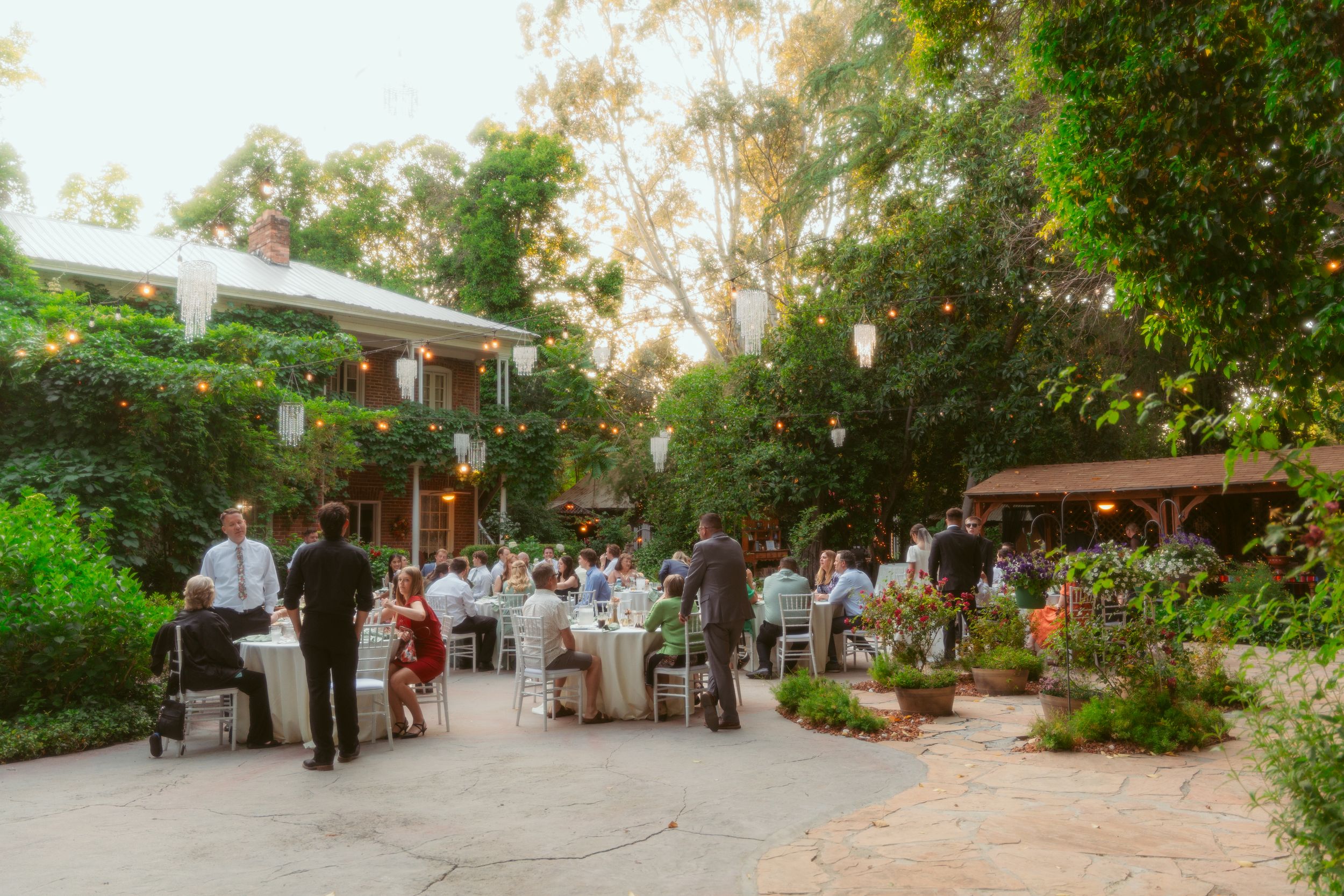 Outdoor garden wedding reception venue with guests dining at tables surrounded by lush greenery at sunset.
