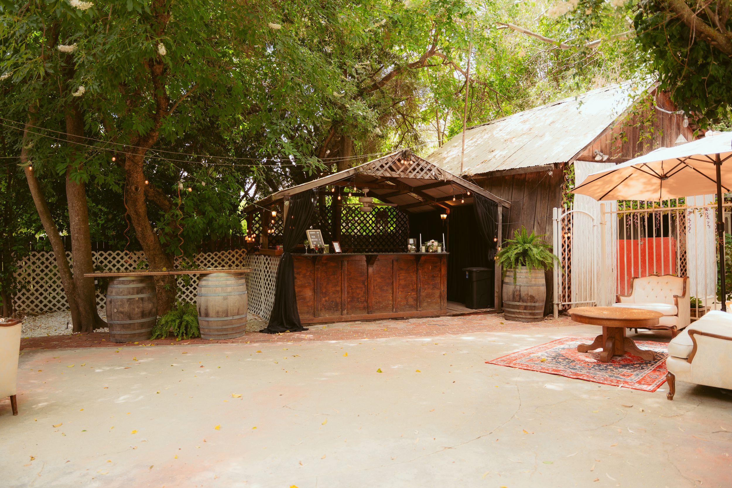 Rustic outdoor wooden bar structure with thatched roof surrounded by lush greenery and picnic tables in a garden setting.