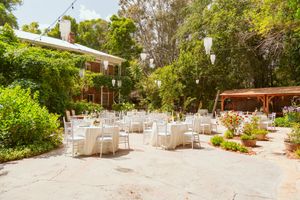 An elegant outdoor garden reception venue with white tables and chairs set up on a sunny patio surrounded by lush greenery.