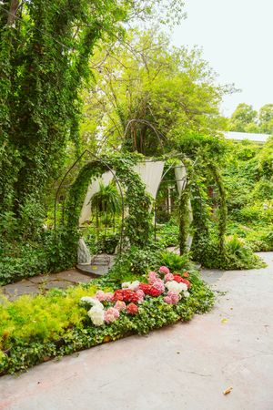 Garden archway decorated with white fabric and pink floral arrangements along a stone pathway in a lush green setting.