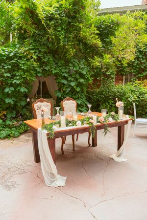 Elegant wedding sweetheart table with vintage chairs and flowing white fabric draped under green foliage outdoors.