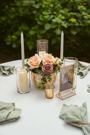 Elegant wedding table centerpiece with white candles, peach and mauve roses, and golden glass votives on white tablecloth.