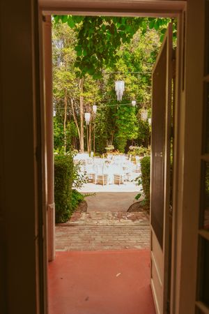 View through open doorway to an outdoor garden wedding reception setup with white chairs and lantern decorations.