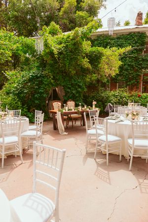 An elegant outdoor wedding reception setup with white Chiavari chairs and tables under lush green foliage.