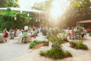 An outdoor garden patio dining area with white tables and chairs surrounded by greenery and glowing sunlight.
