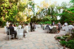 Outdoor garden reception with guests seated at round tables on a stone patio surrounded by lush greenery at dusk.