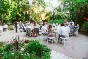 Outdoor garden wedding reception with guests seated at round tables under lush greenery and string lights.