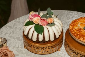 A round bundt cake decorated with white frosting and pink roses sits on an ornate copper cake stand.