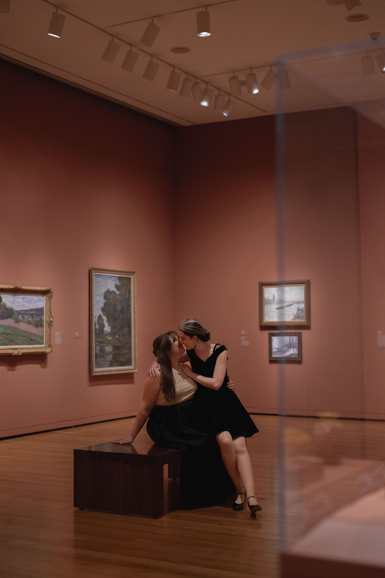 Two people in black outfits share an intimate moment while sitting on a bench in an art gallery with warm terracotta walls.
