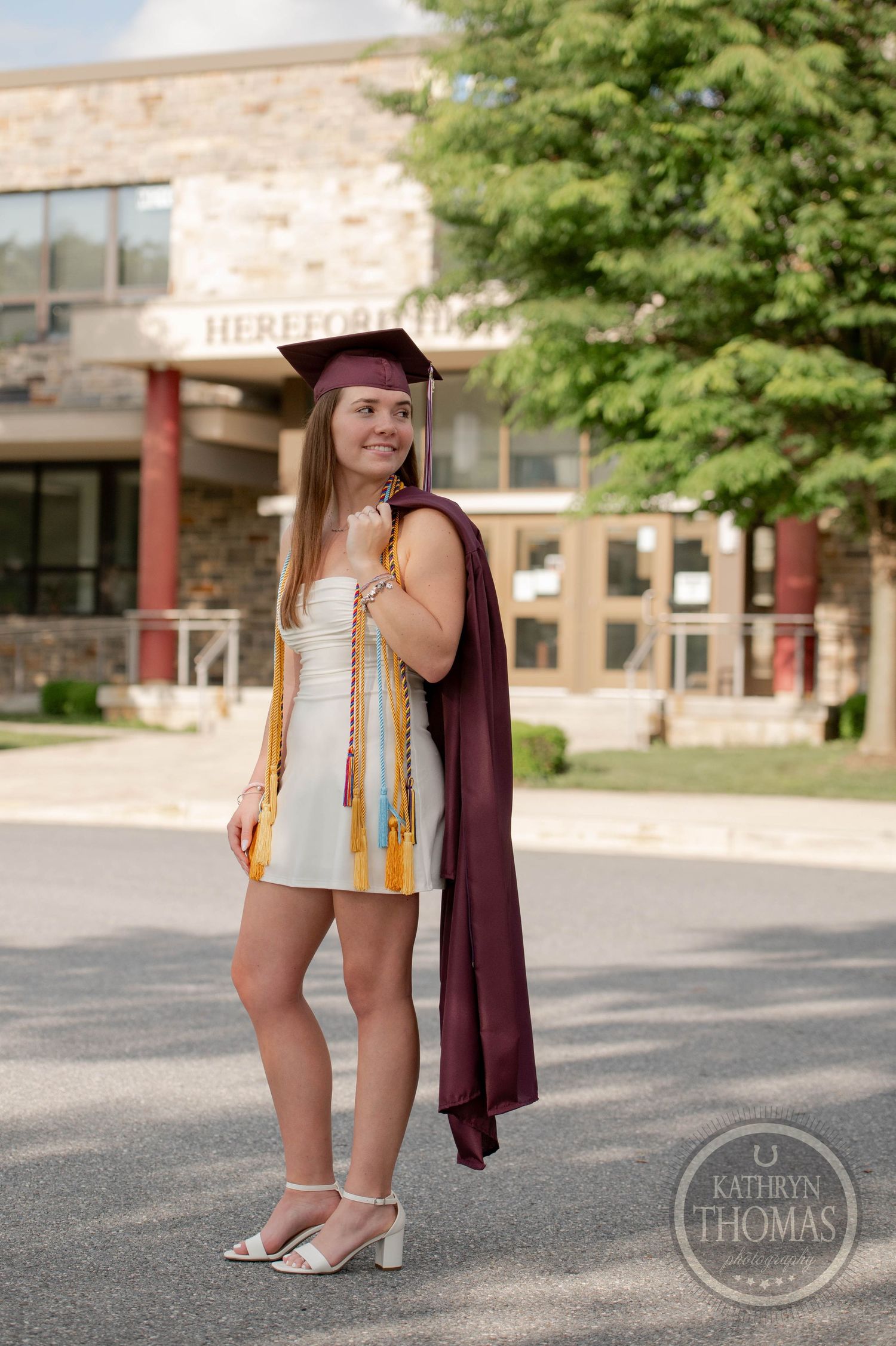 Graduate wearing maroon cap and gown with gold honors cords poses outside brick university building on sunny day.