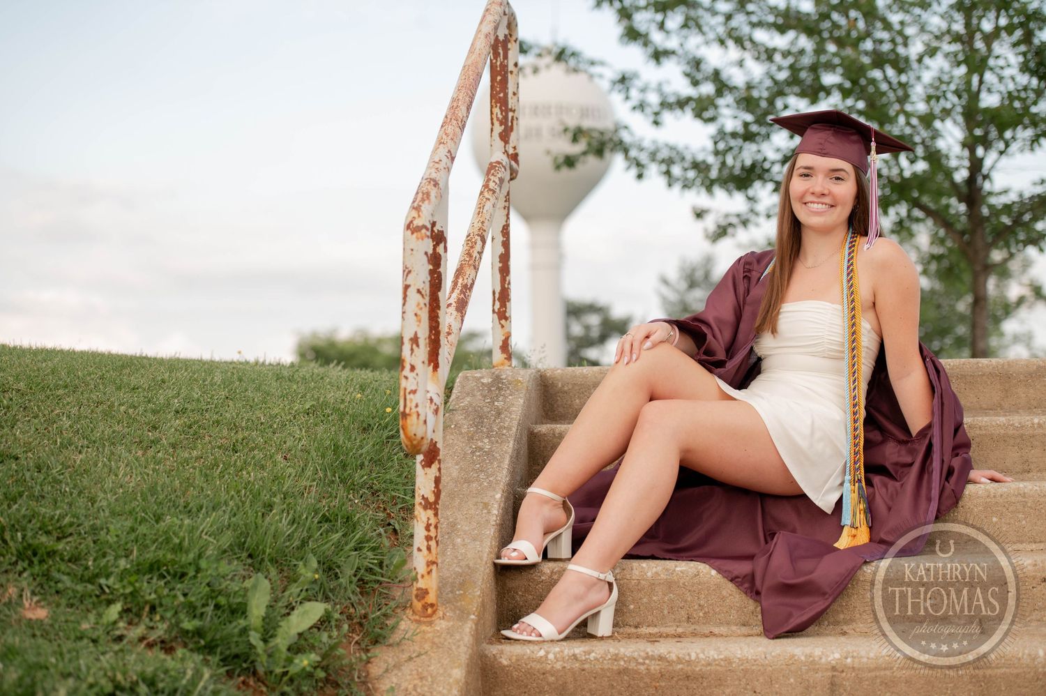 Graduate in maroon cap and gown sits on concrete steps outdoors with metallic decorative railing.