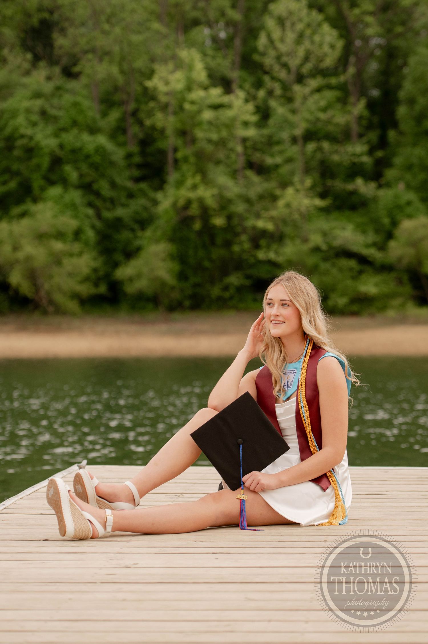 Figure sits peacefully on a wooden dock by a serene lake surrounded by lush green forest on a summer day.