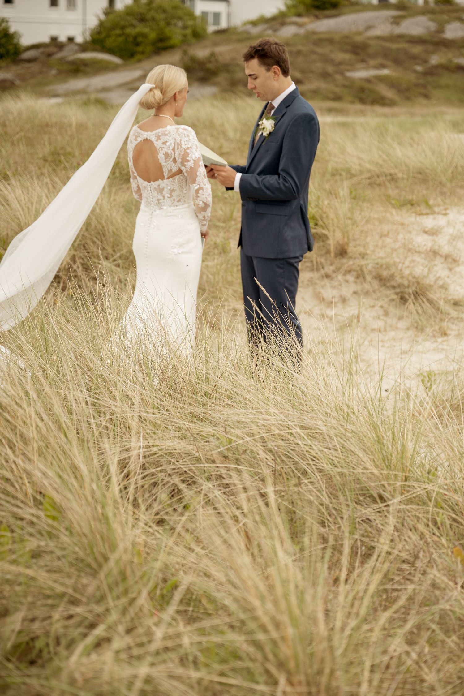 A romantic wedding couple embraces in a desert field at sunset, with the bride wearing a flowing lace gown.