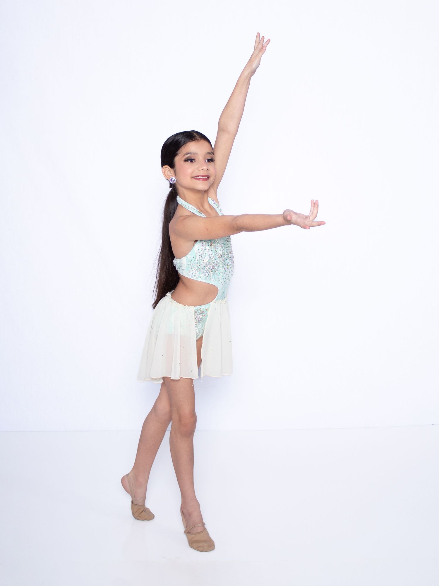 Young dancer in sparkly white dance costume striking graceful pose with arms outstretched against white background.