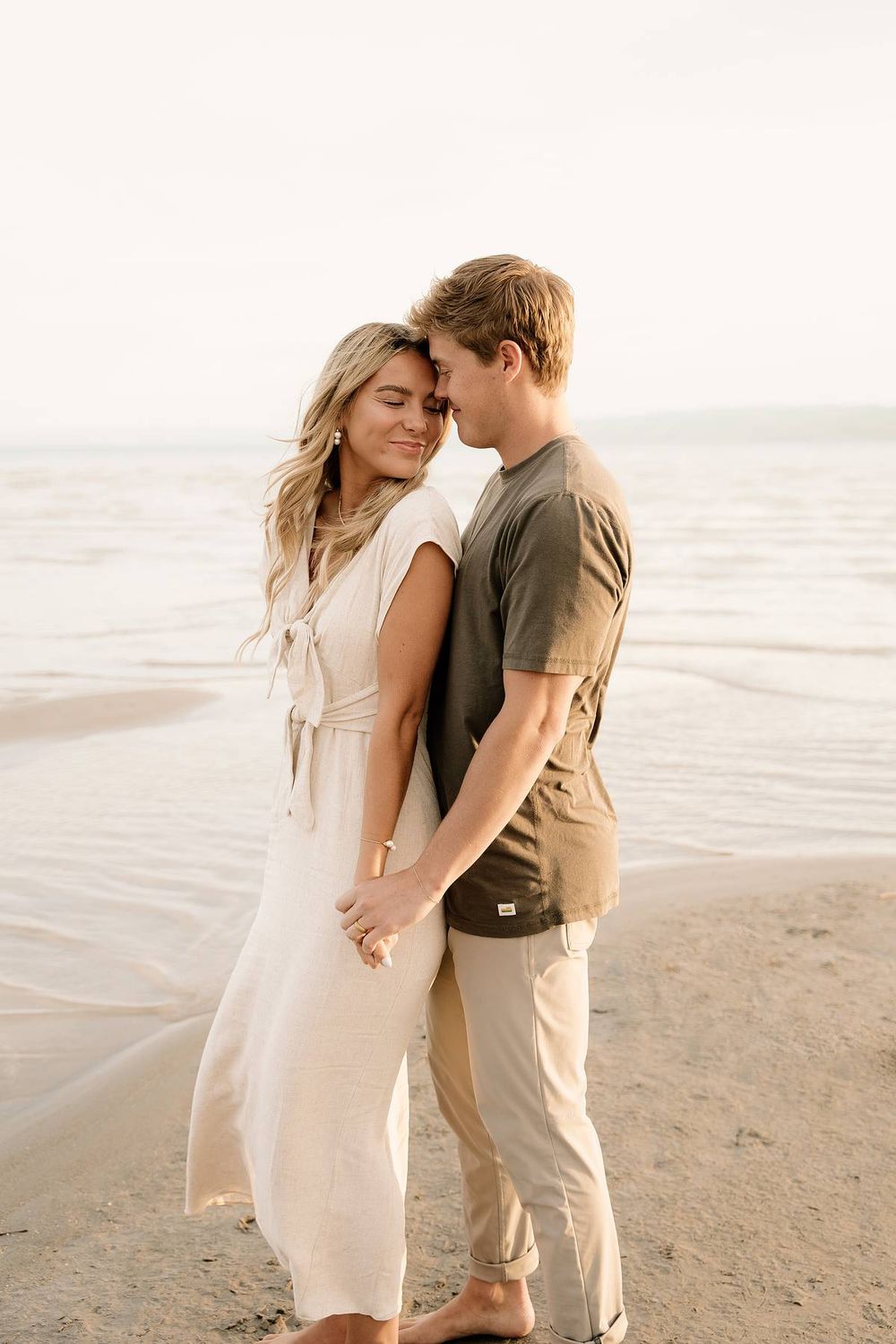 Intimate moment between couple wearing neutral colors embracing on peaceful beach at sunset.