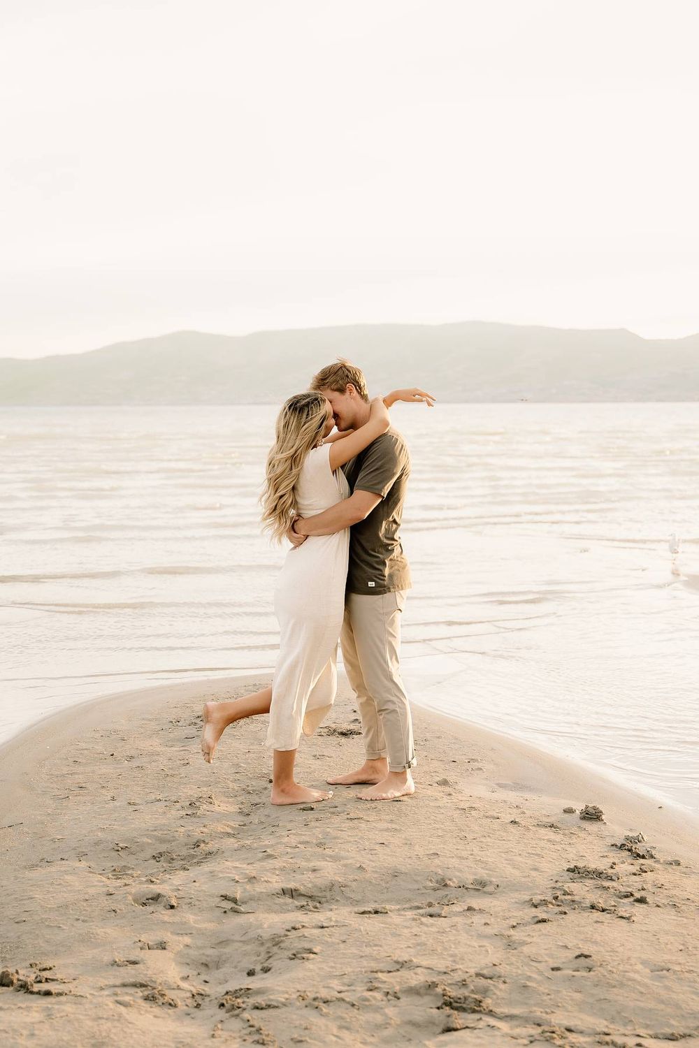 Couple in white outfits sharing a romantic embrace on a beach with mountains in the background.