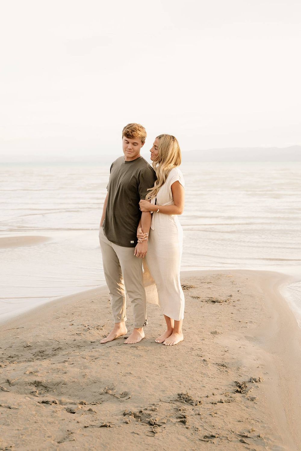 A romantic couple stands barefoot on a beach together during a serene coastal photoshoot.