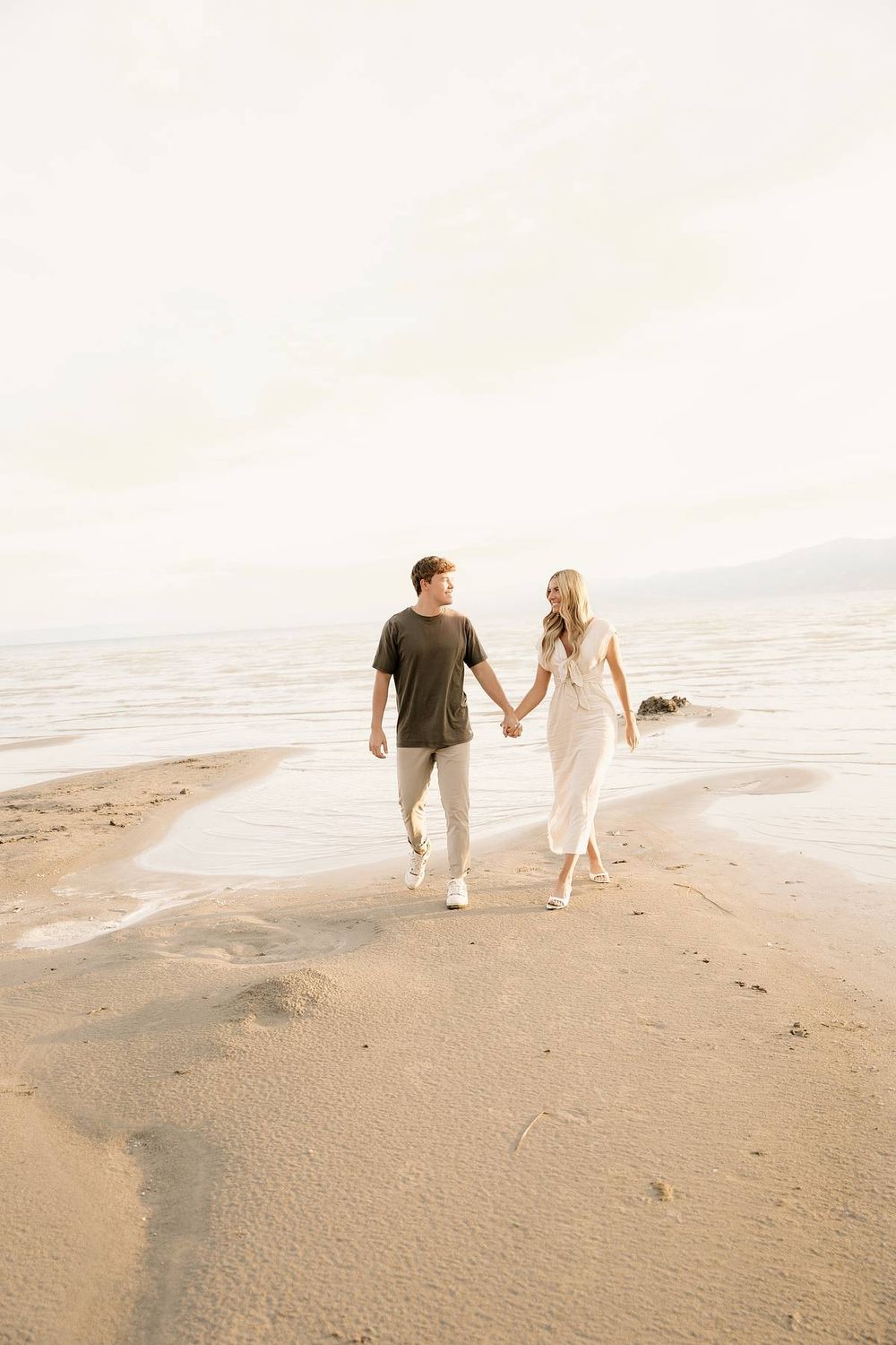 Couple walking hand in hand along shoreline during golden hour.