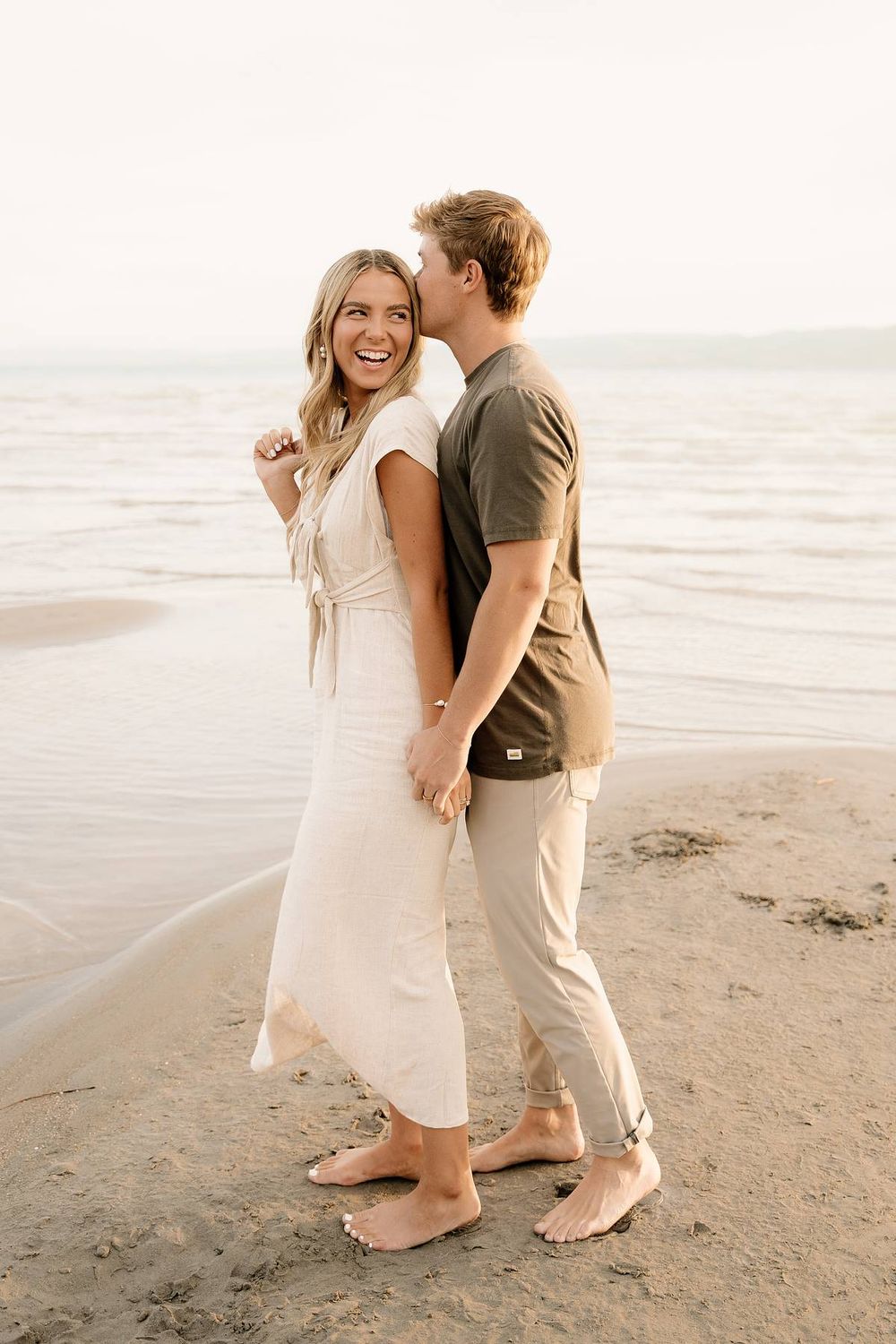 Couple embracing and laughing together on a beach at sunset wearing casual white and brown outfits.