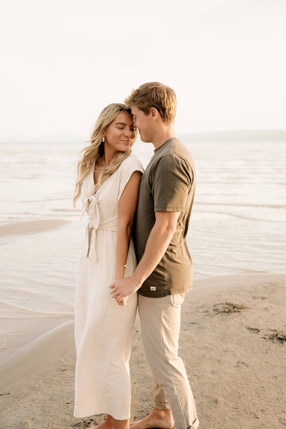 A tender moment between a couple on a sunlit beach wearing light colored clothing.