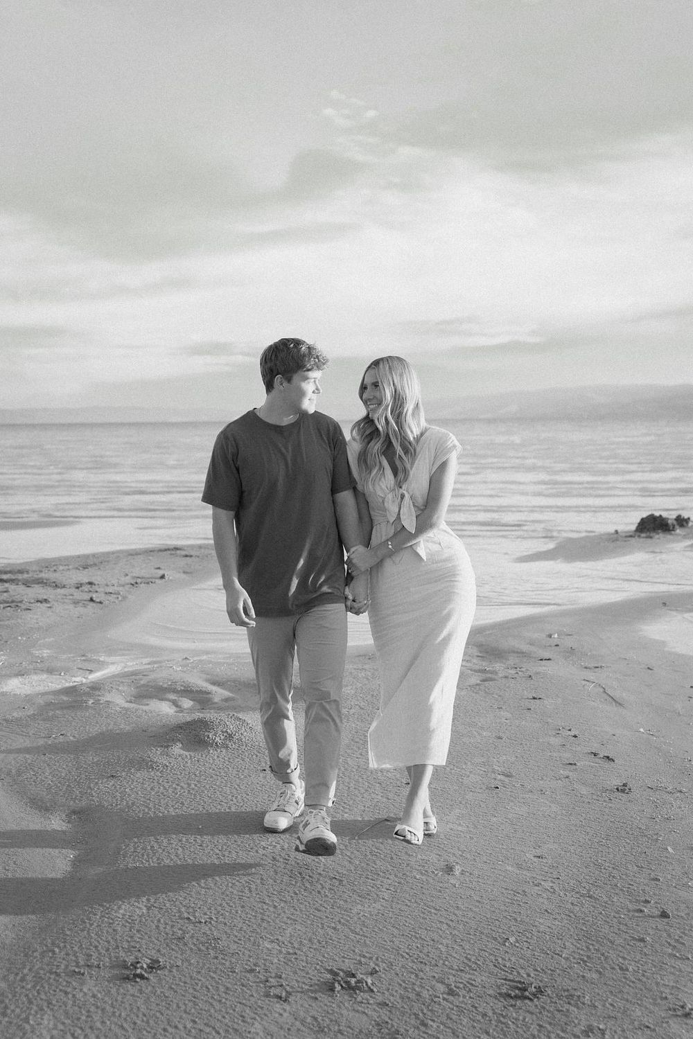 Couple walking hand in hand along a peaceful beach shore in black and white.