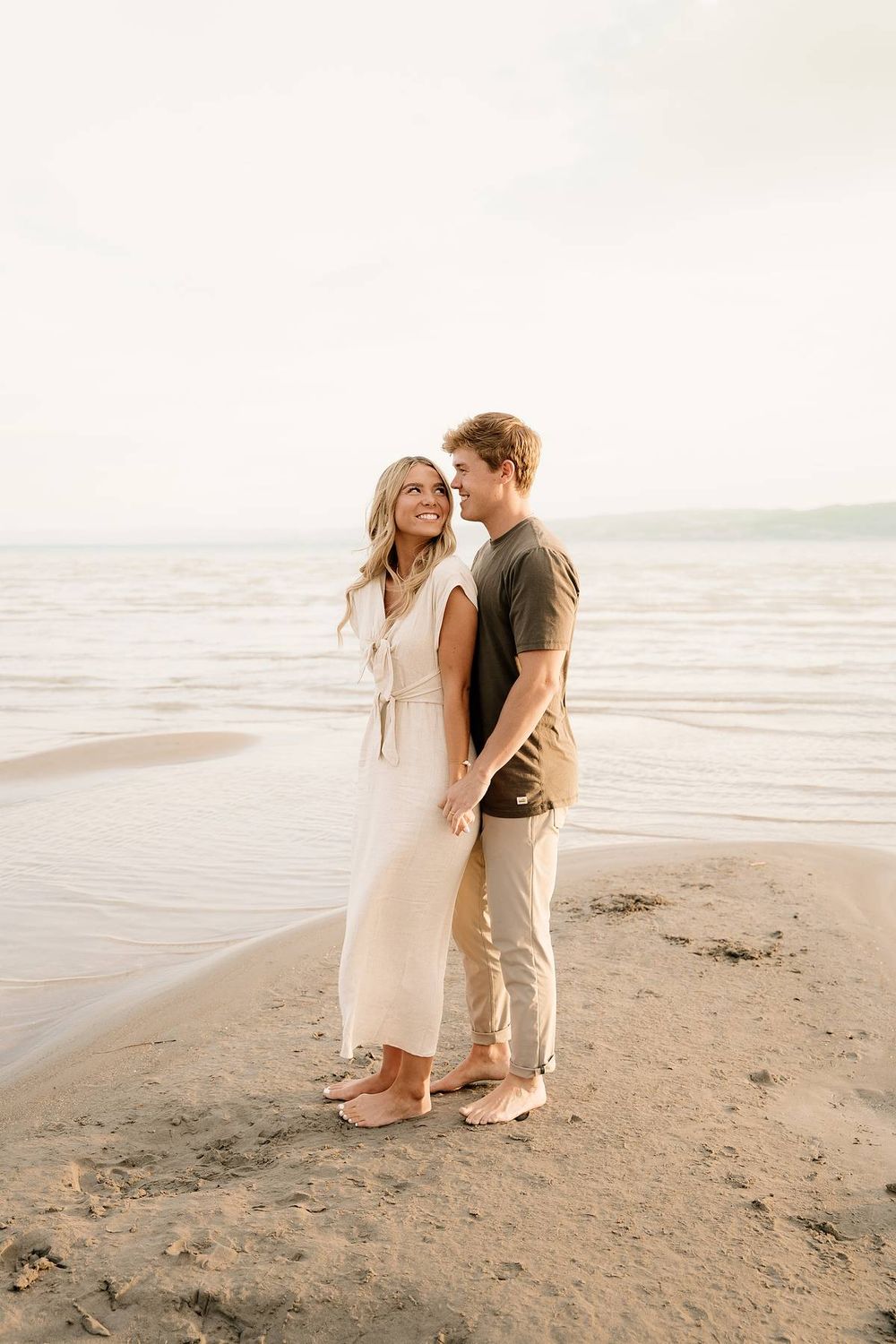 A romantic couple stands together on a sandy beach at sunset in casual summer attire.
