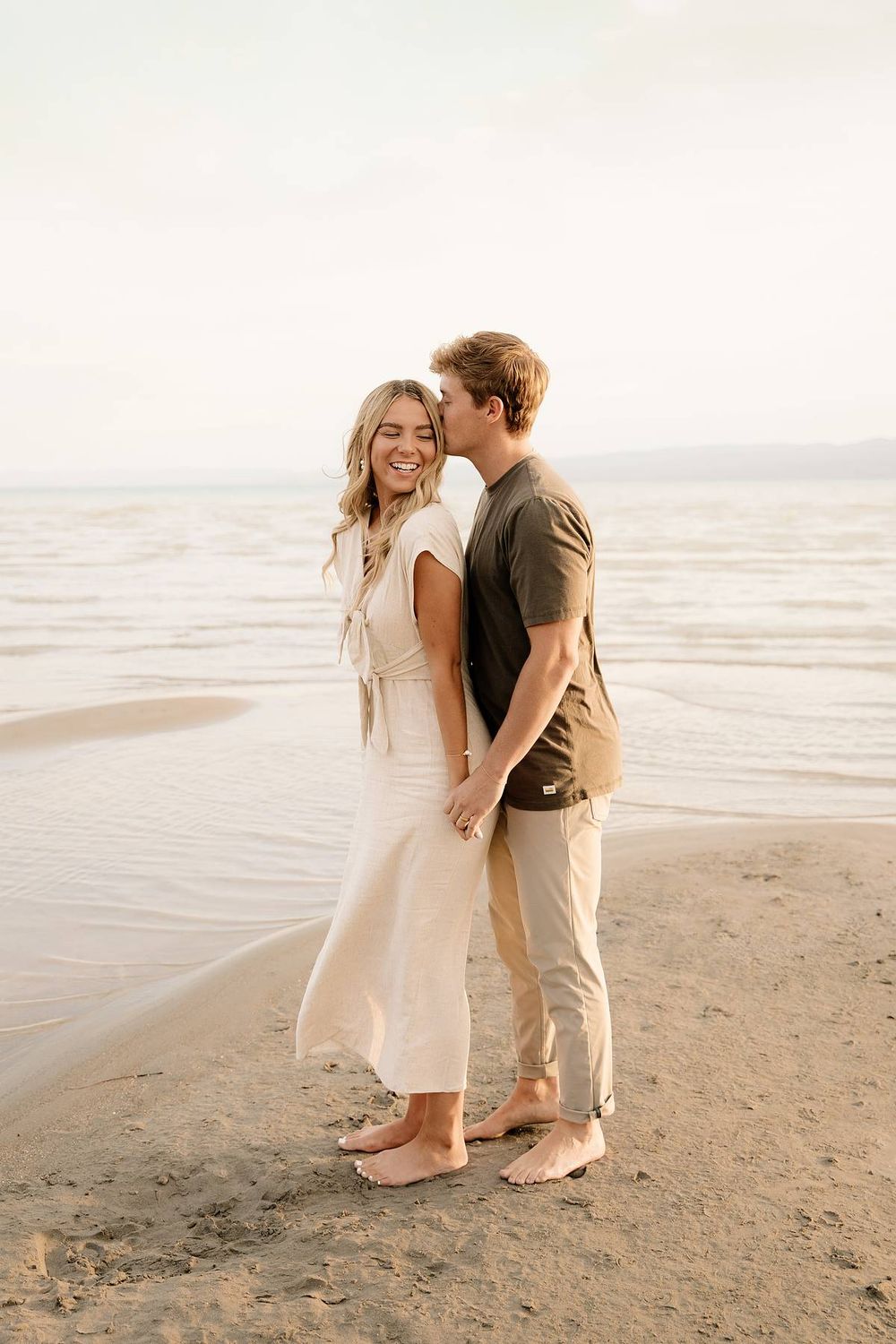 A romantic couple embraces on a serene beach at sunset while wearing casual neutral-toned outfits.