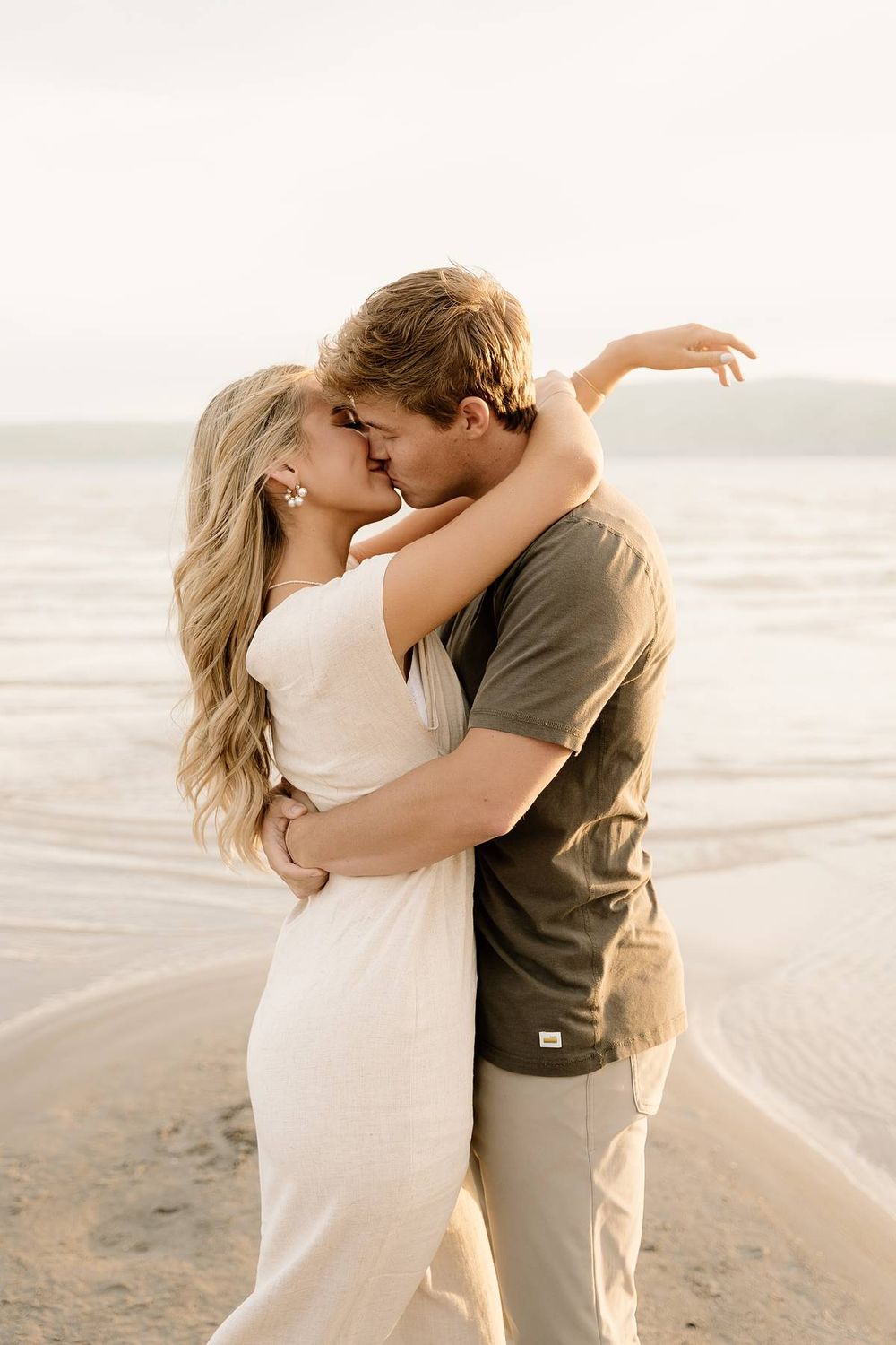 A romantic couple embraces and kisses on a serene beach at sunset with waves in the background.