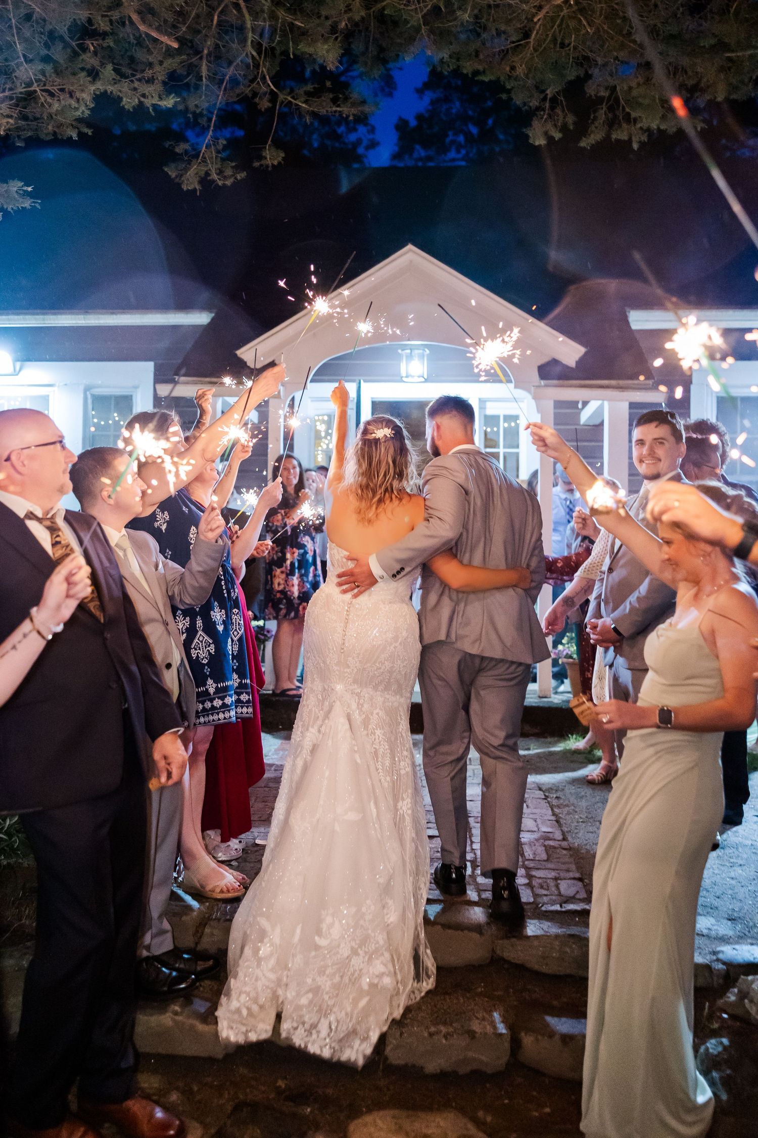 Wedding guests hold sparklers creating a magical tunnel of light as the bride and groom exit their reception at night.