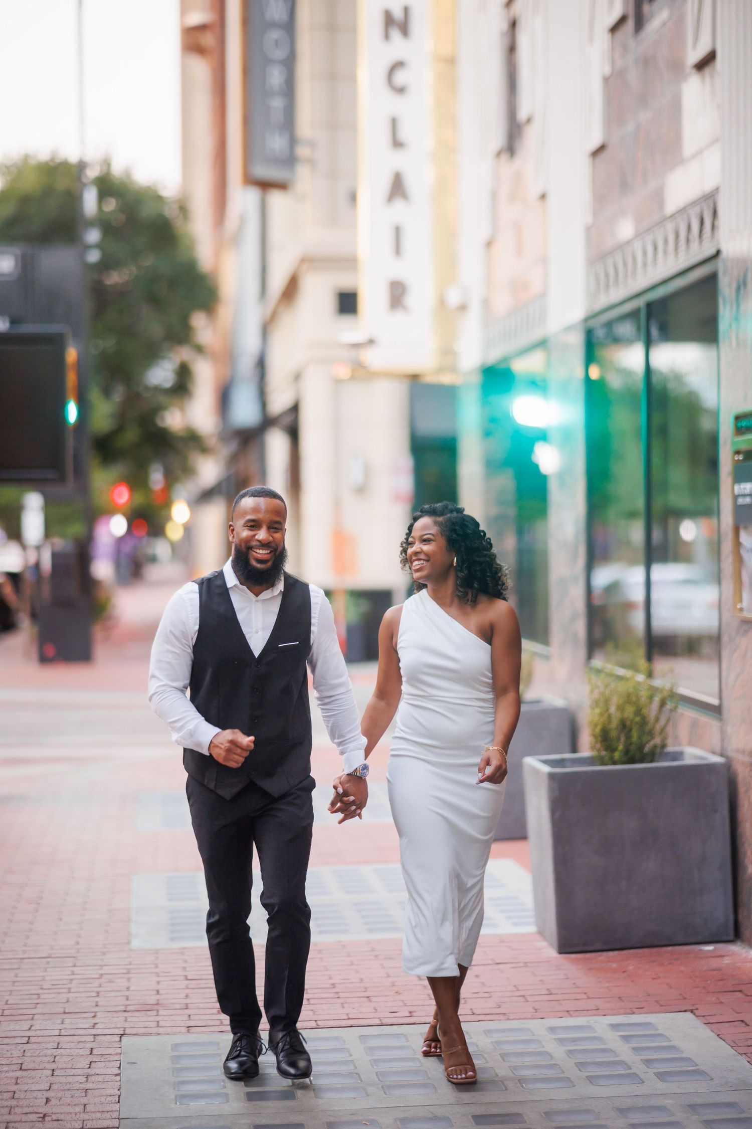 A woman in a white dress and man in a black vest and suit walks together while laughing on a Dallas sidewalk during sunset.