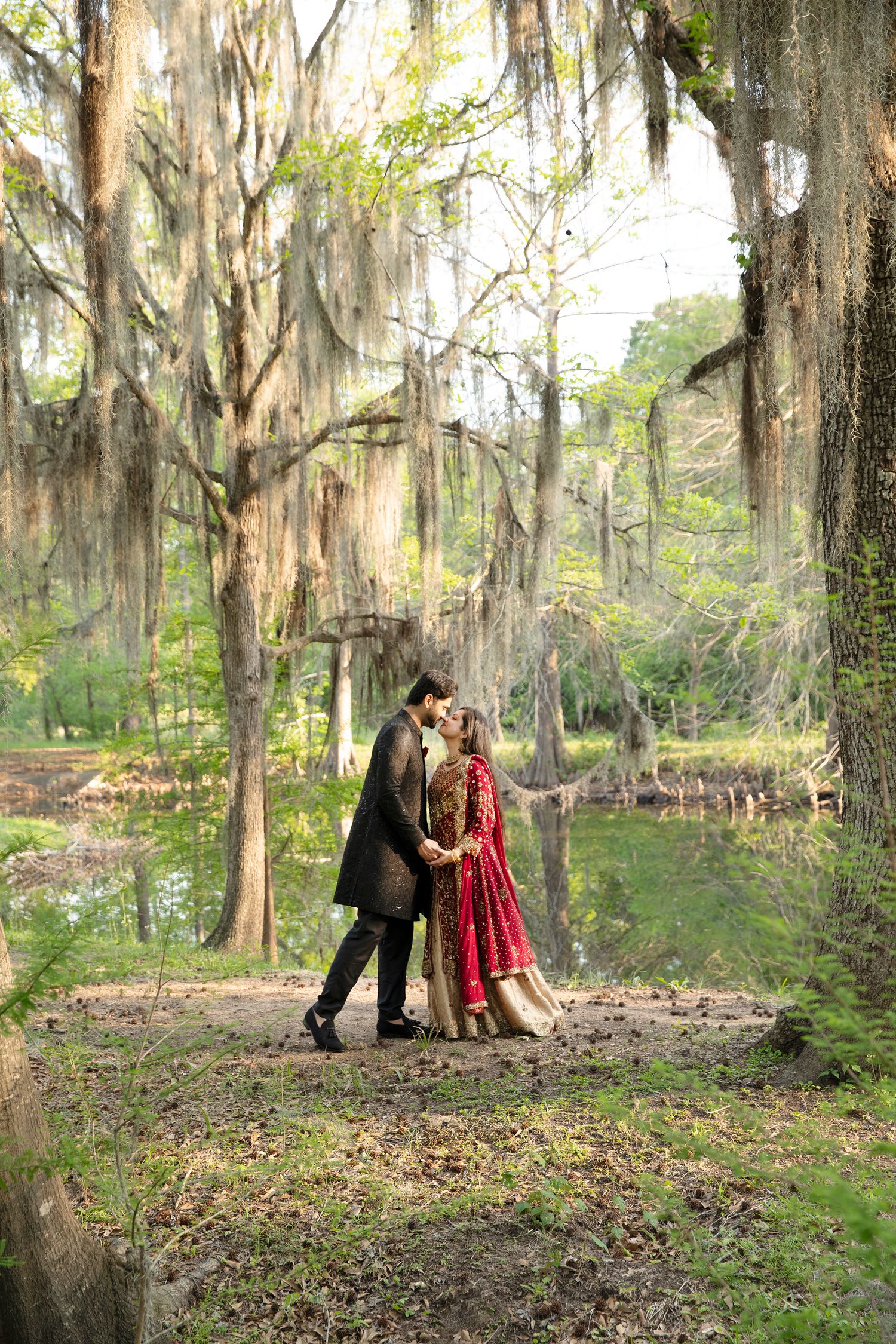 A couple embraces under moss-draped oak trees beside a tranquil pond in a romantic Southern garden setting.