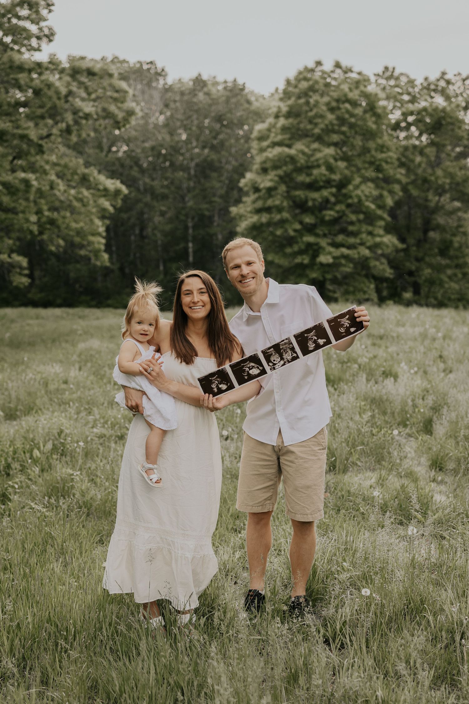 A family stands together in a grassy meadow holding ultrasound photos while announcing a pregnancy.