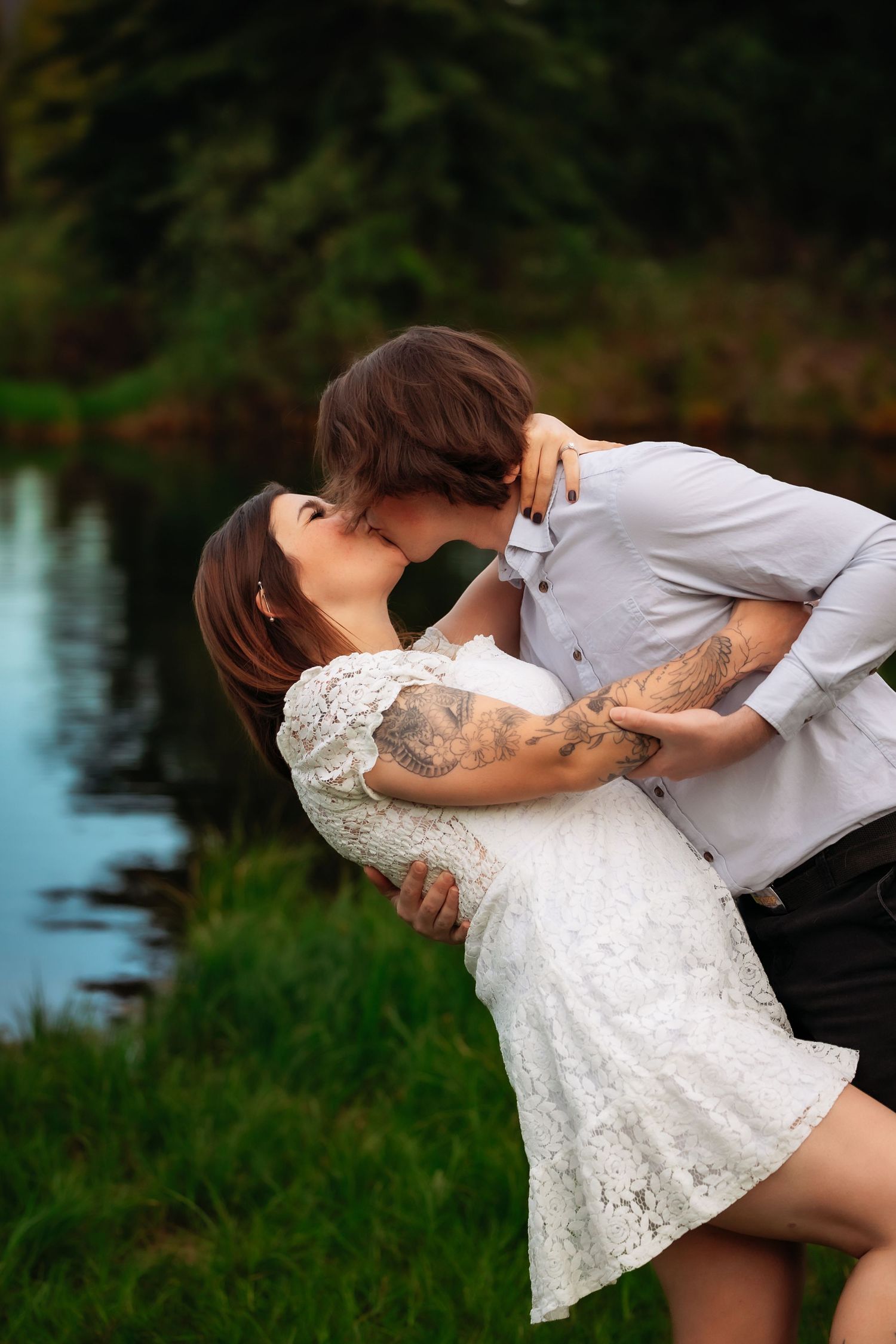 A romantic couple embraces in a dip pose by a lake, with the individual in a white lace dress and the other in gray.