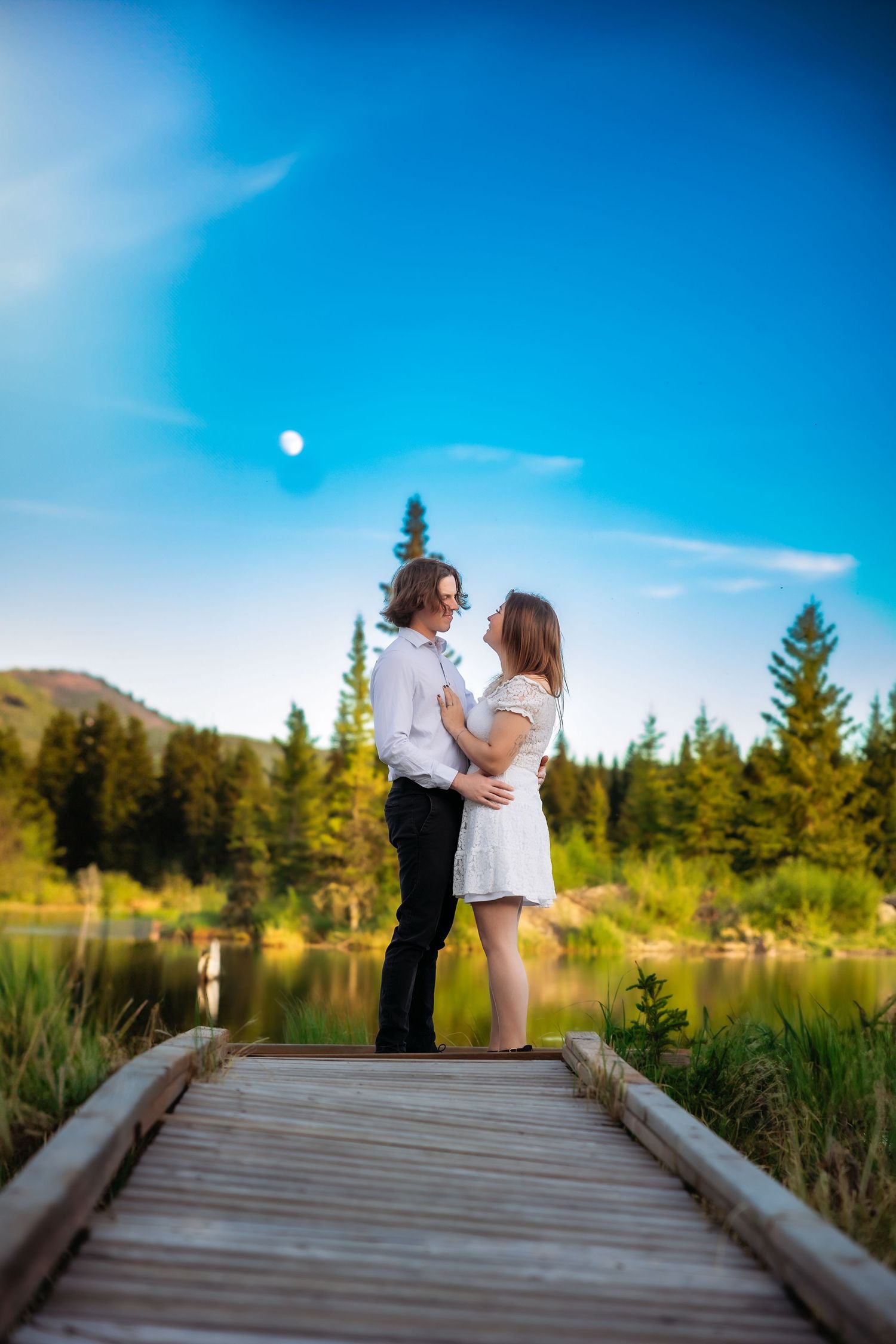 A romantic moment captured on a wooden dock overlooking a serene mountain lake with evergreen trees in the background.