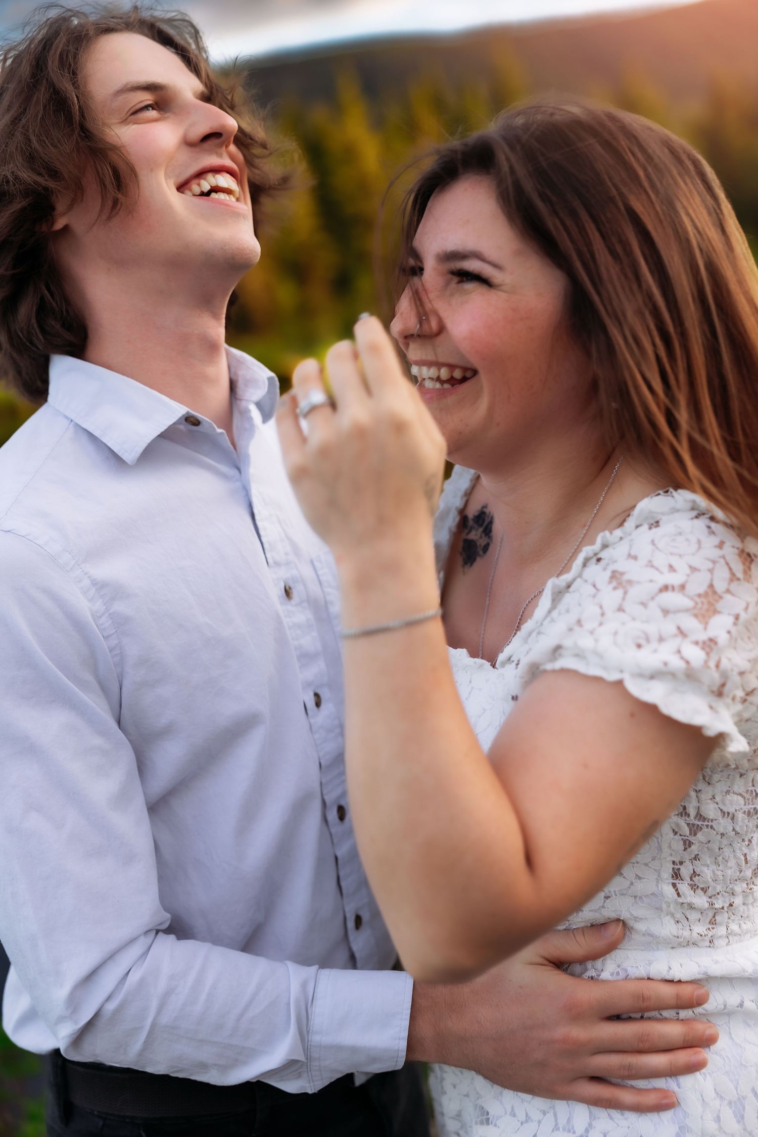 A couple laughs together outdoors, with the person in white lace and their partner in a light button-down shirt at sunset.