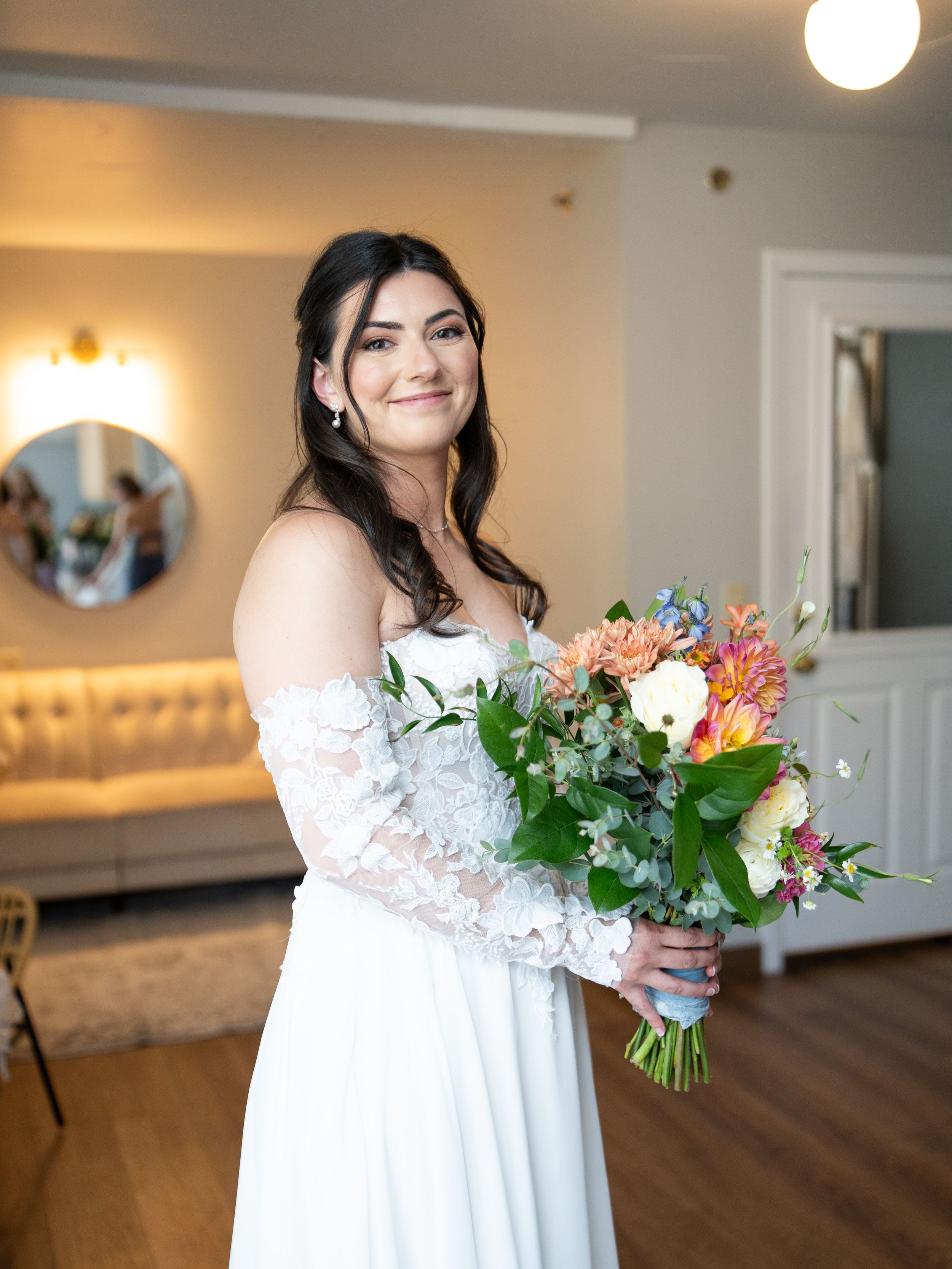 A bride in a lace off-shoulder wedding dress smiles while holding a vibrant bouquet with peach and pink blooms at Tannenbaum.