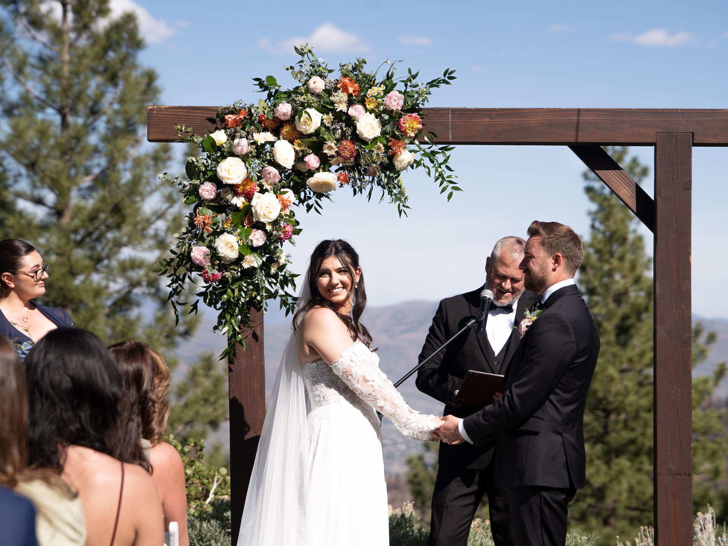 Outdoor wedding ceremony under wooden arch decorated with peach and white floral arrangements against mountain backdrop.