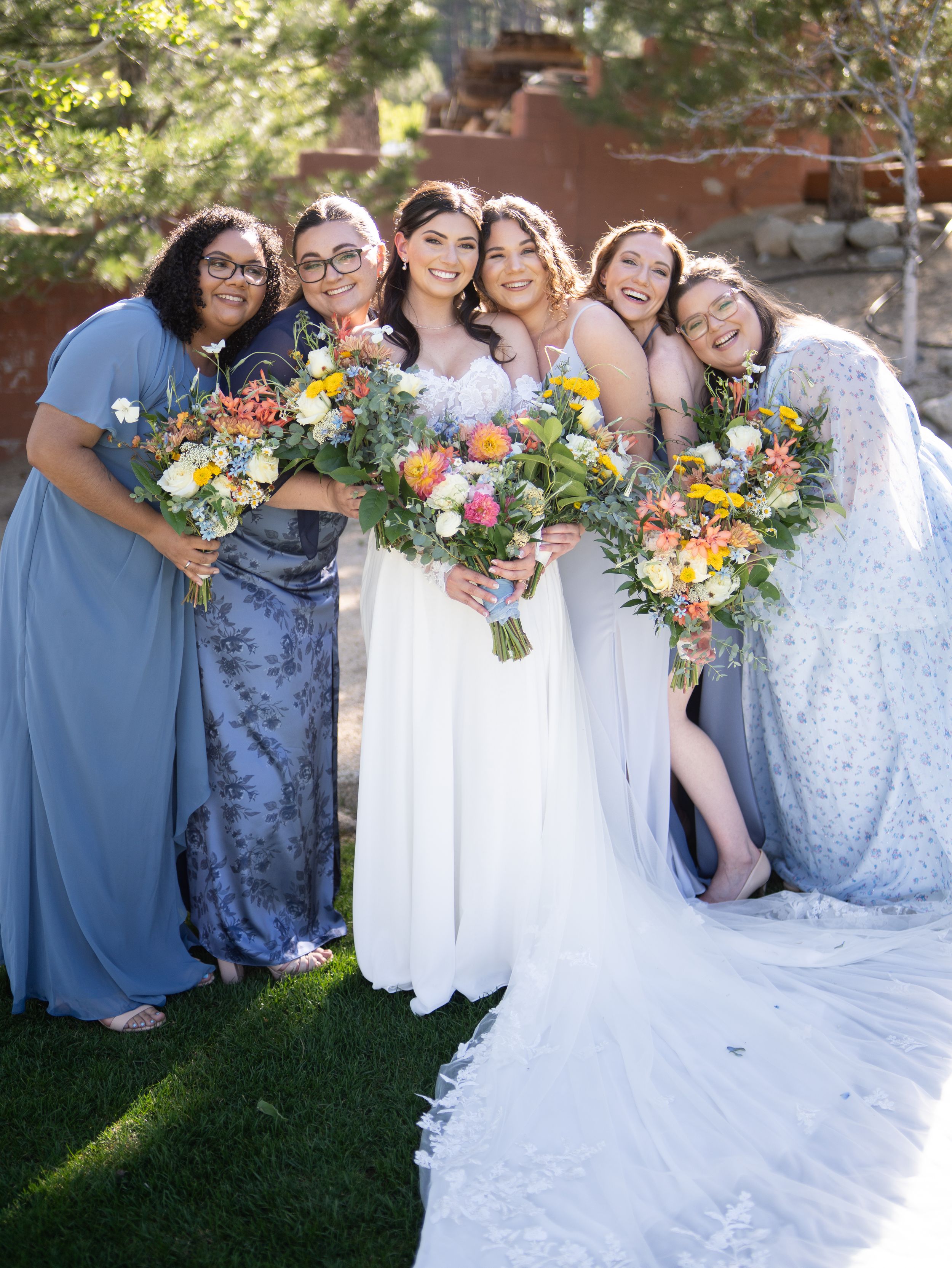 A bridal party wearing blue and floral dresses poses outdoors while holding vibrant mixed flower bouquets on a sunny day.