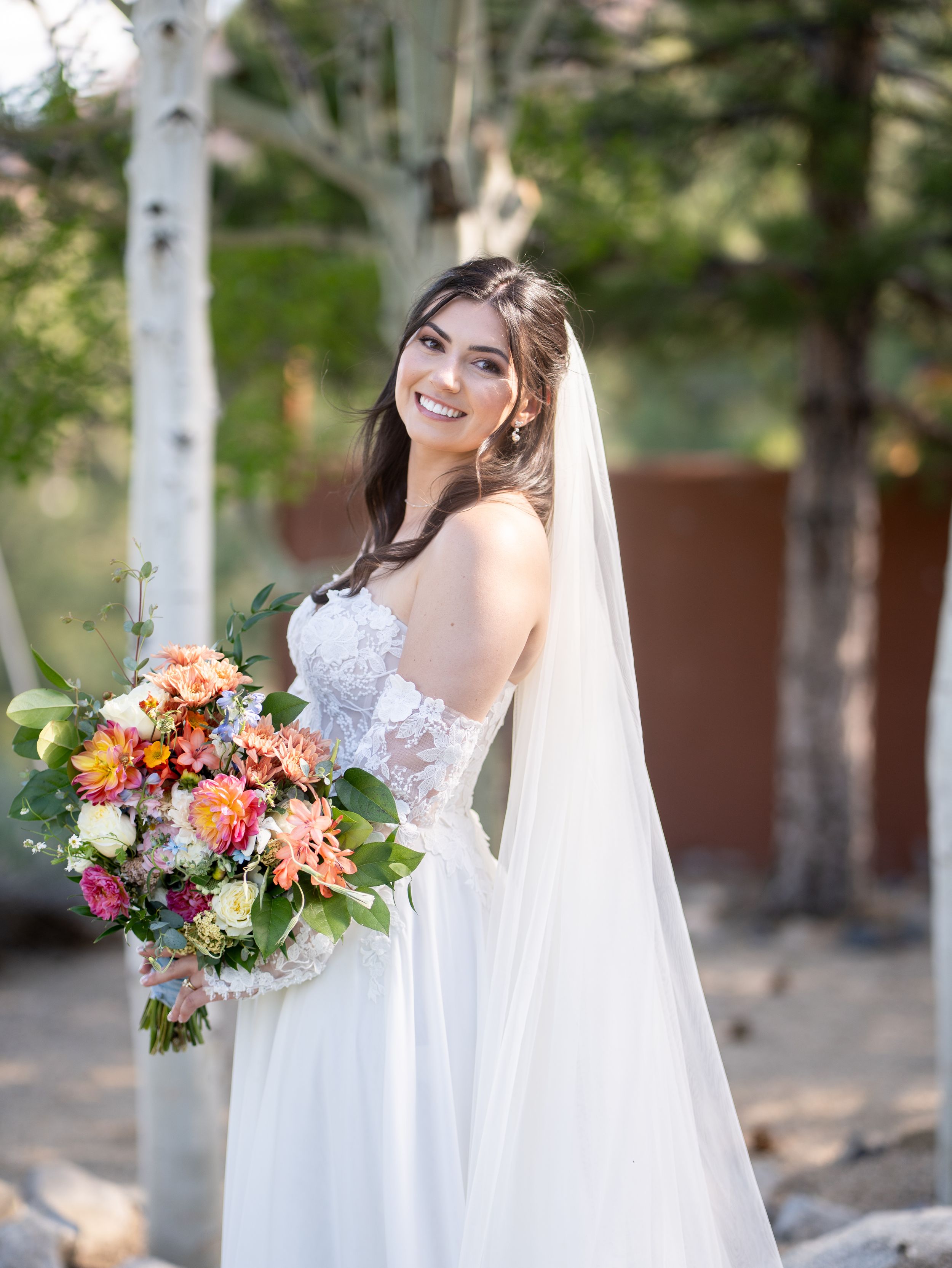 A bride in a white lace wedding dress poses outdoors with a vibrant coral and pink floral bouquet next to birch trees.