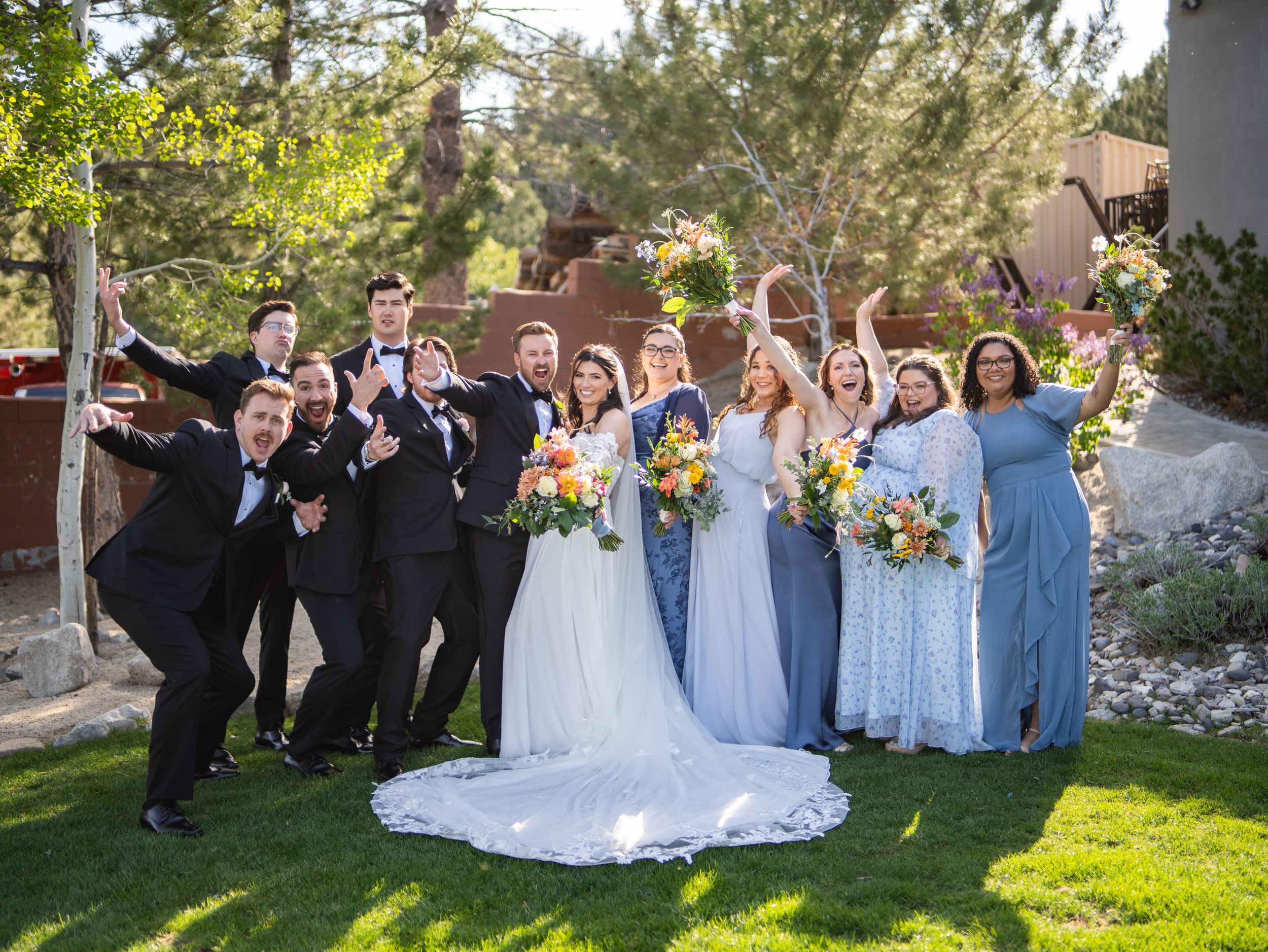 A wedding party poses outdoors with groomsmen in black suits and bridesmaids in light blue dresses holding yellow bouquets.