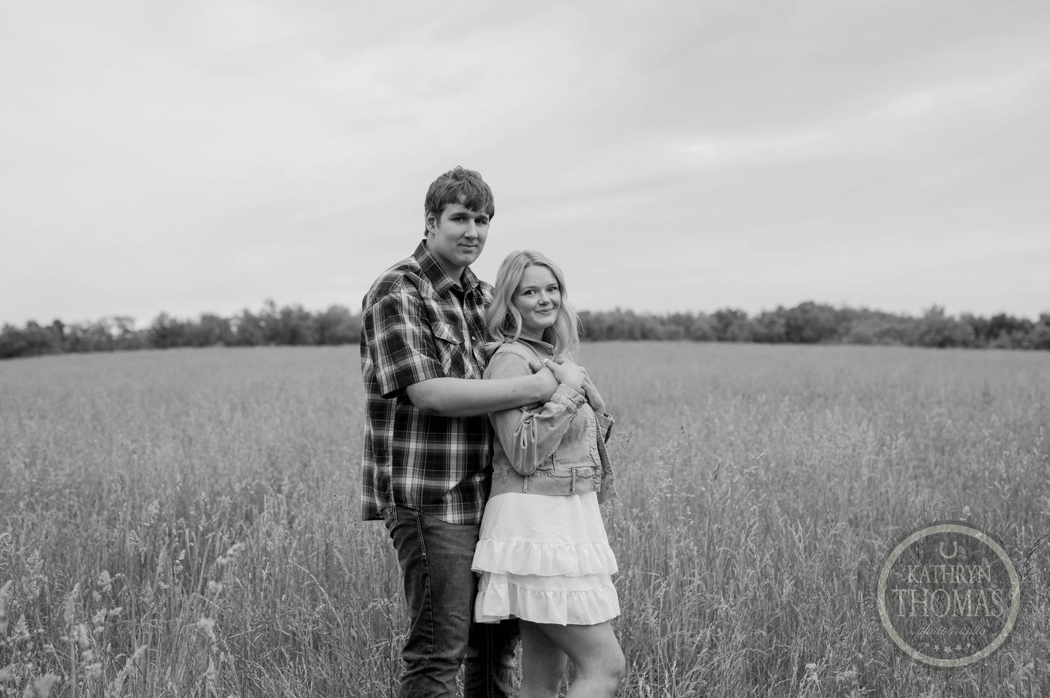 A couple stands embracing in a rustic field setting captured in black and white photography.