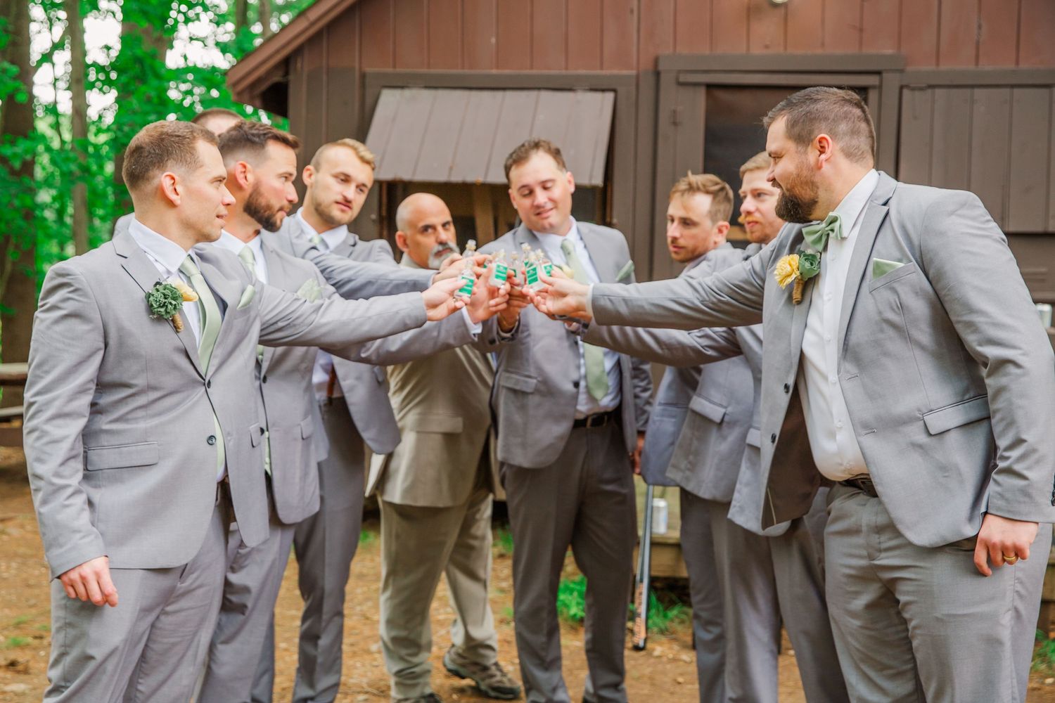 Groomsmen in matching grey suits toast and clink glasses together in celebration at outdoor wedding venue.