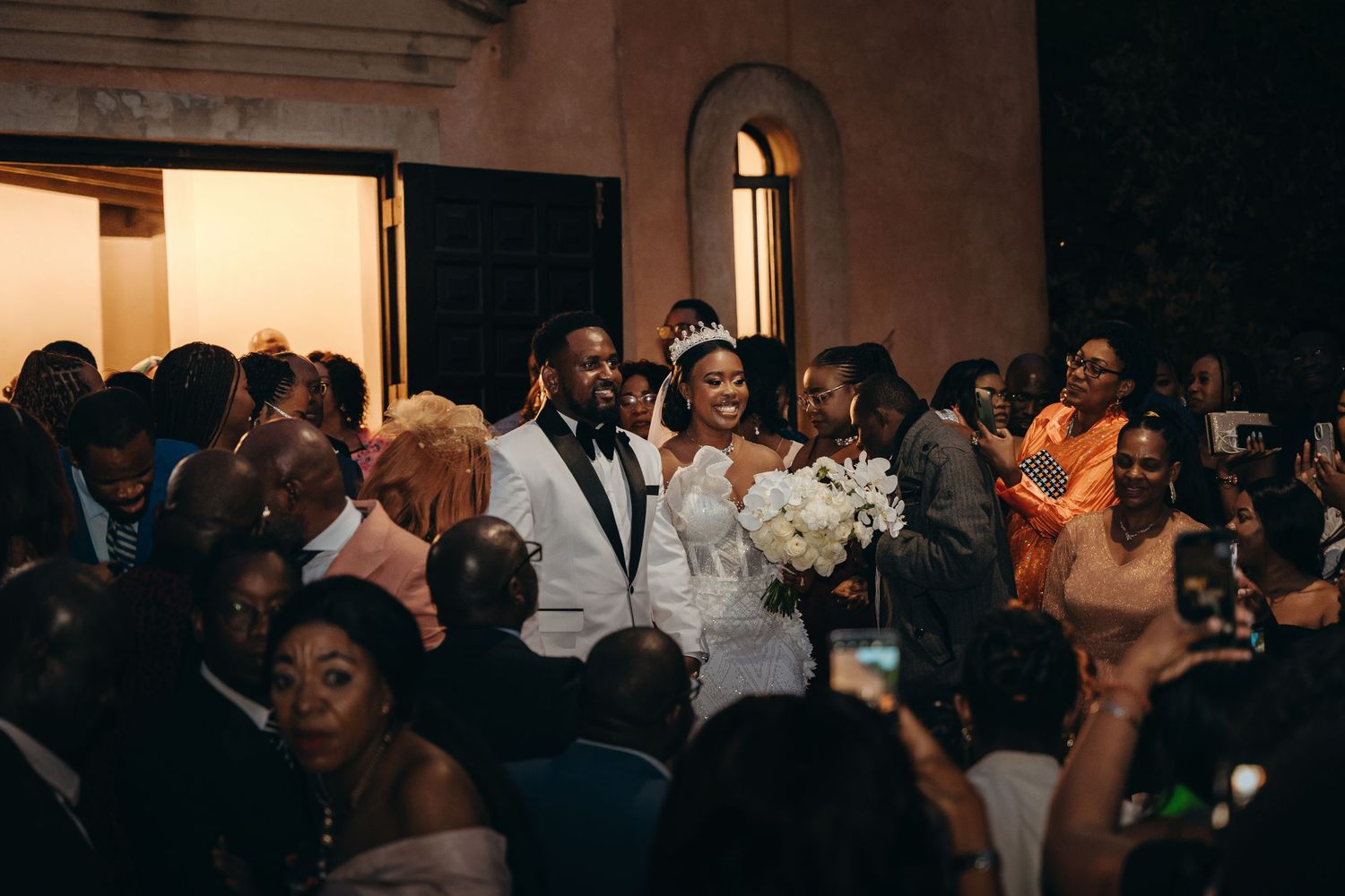 Artistic wedding photo of a couple in formal attire posing against a salmon colored wall with architectural details.
