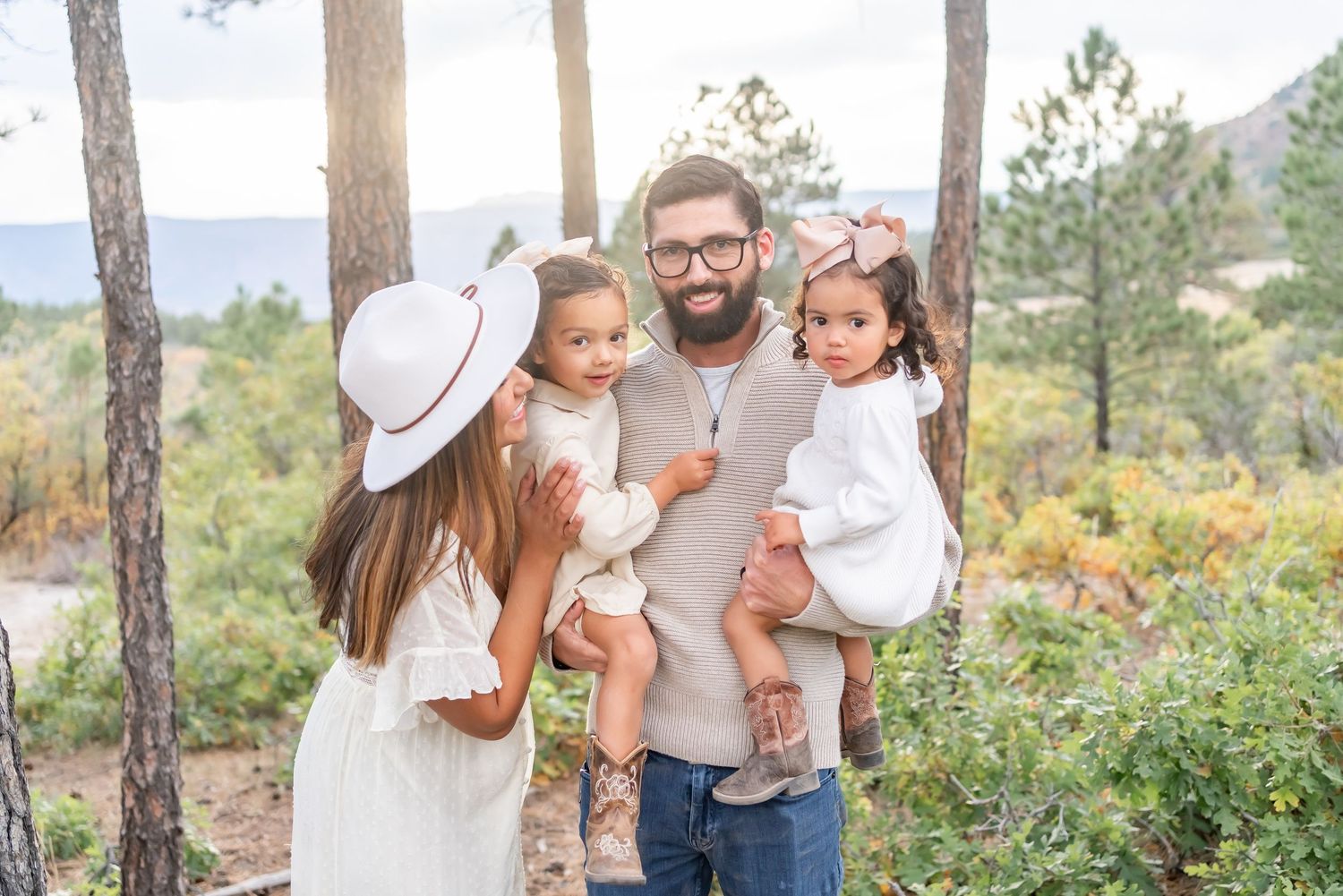 A family wearing white and neutral tones stands together in a scenic mountain forest landscape during golden hour.