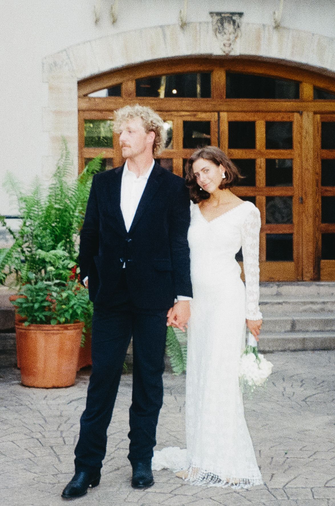 A couple shares a romantic moment under a clear umbrella in a garden archway with red terracotta tiles.