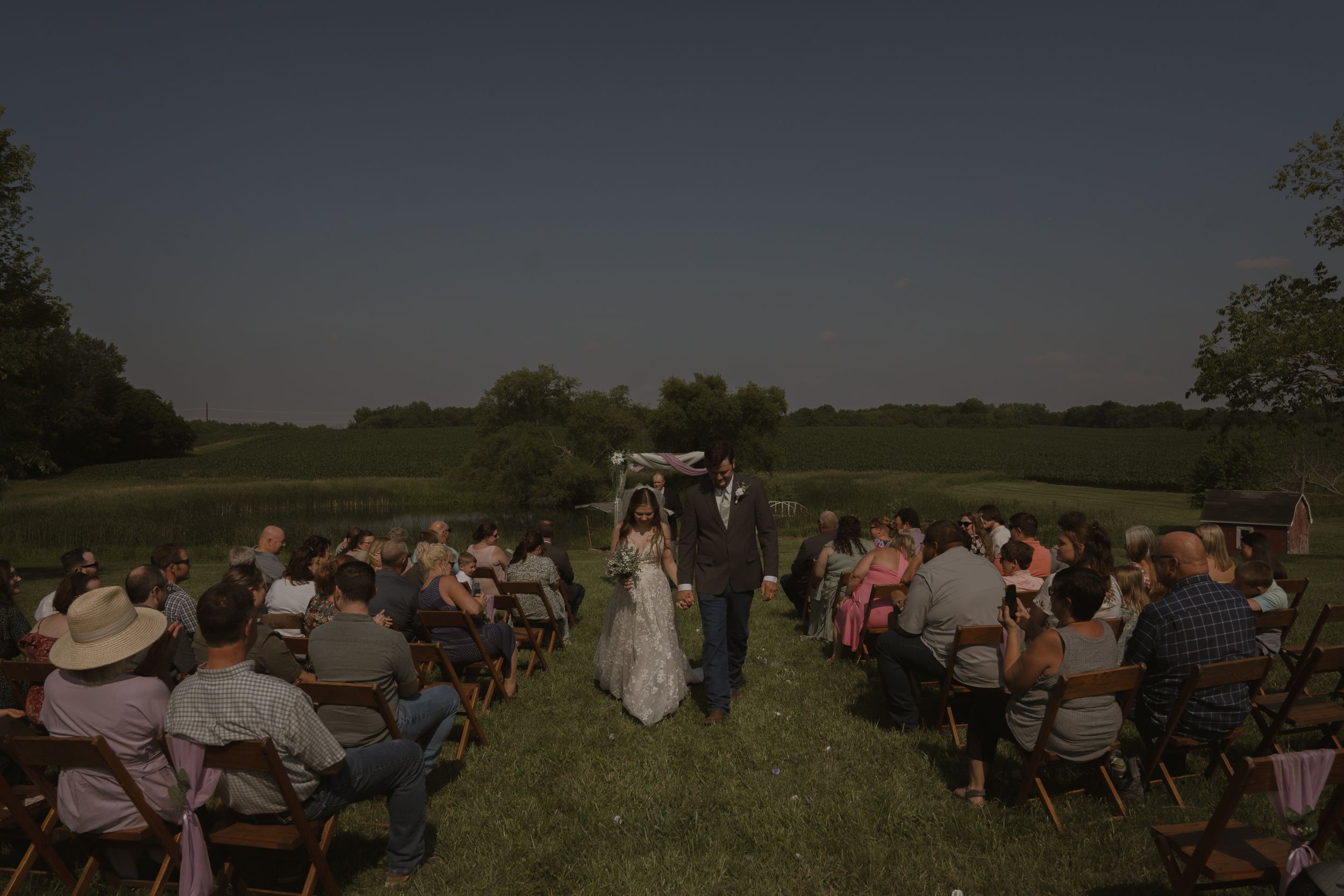 Wedding guests seated in wooden chairs for an outdoor ceremony in a scenic field.