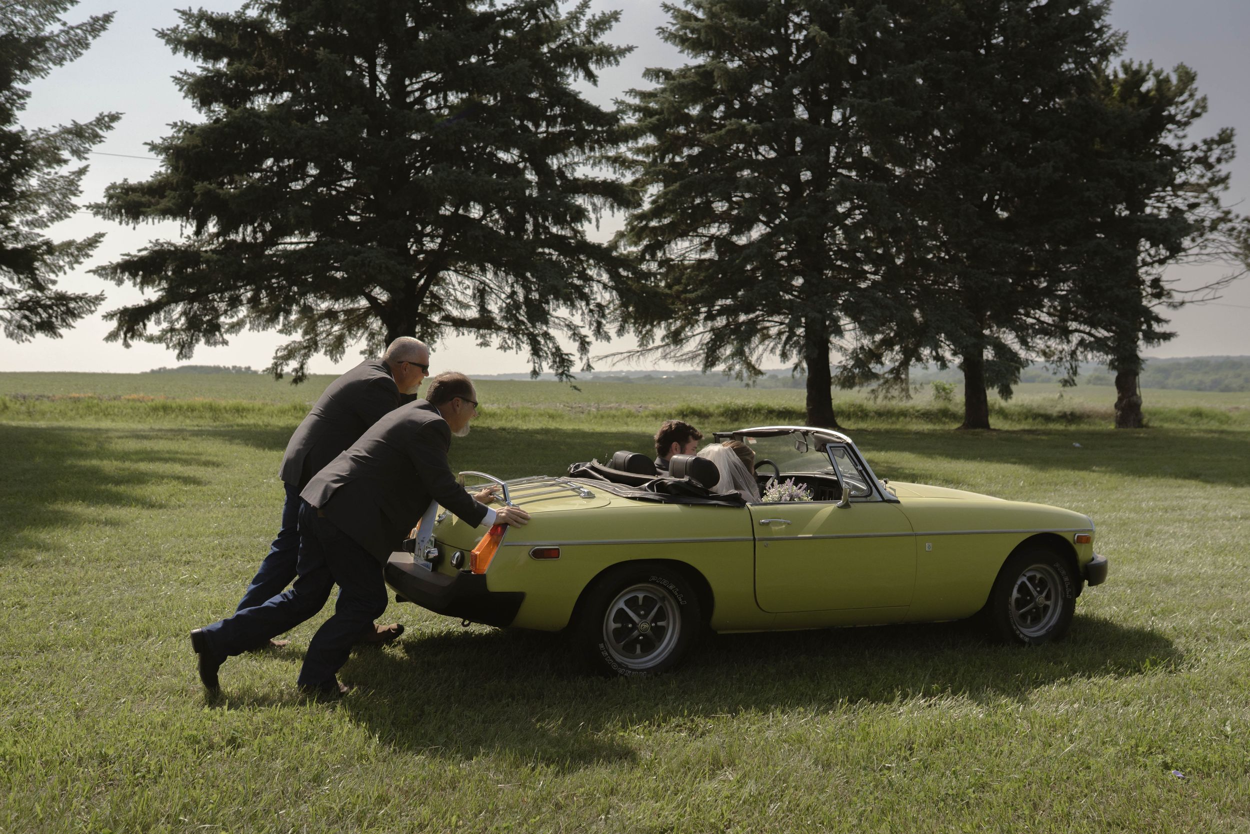 Someone pushes a classic yellow convertible car across a grassy field.