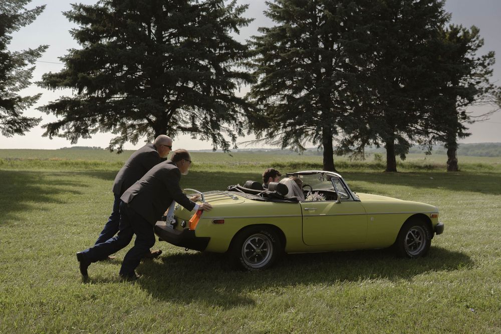 Someone pushes a classic yellow convertible car across a grassy field.